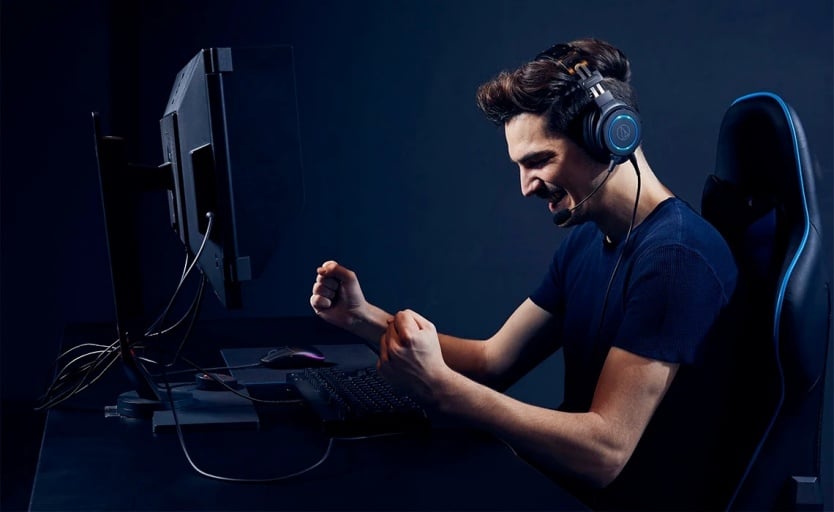 mężczyzna siedzi przed komputerem i unosi ręce w geście zwycięstwa, na uszach ma słuchawki Audio-Technica ATH-G1