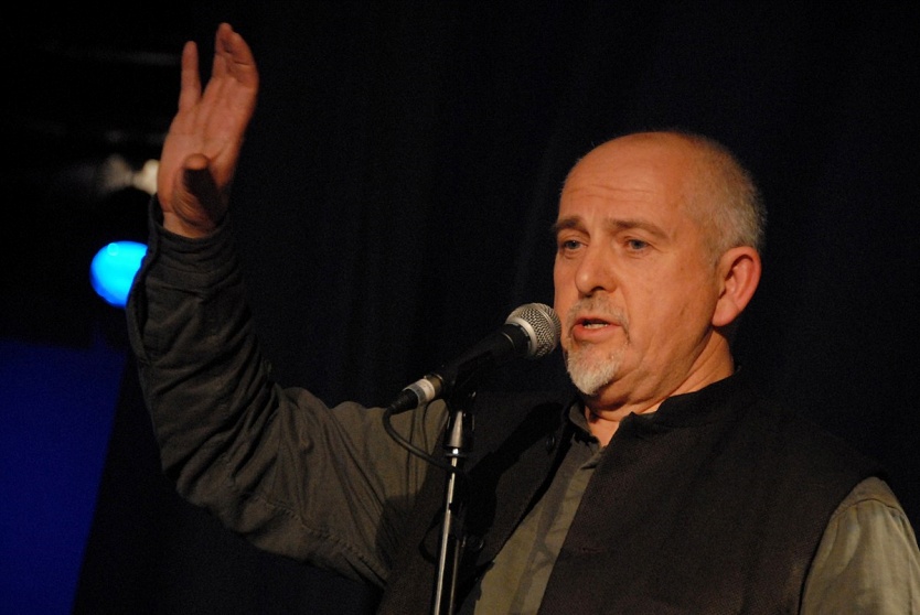 Peter Gabriel stoi przed mikrofonem, prawą rękę ma uniesioną do góry