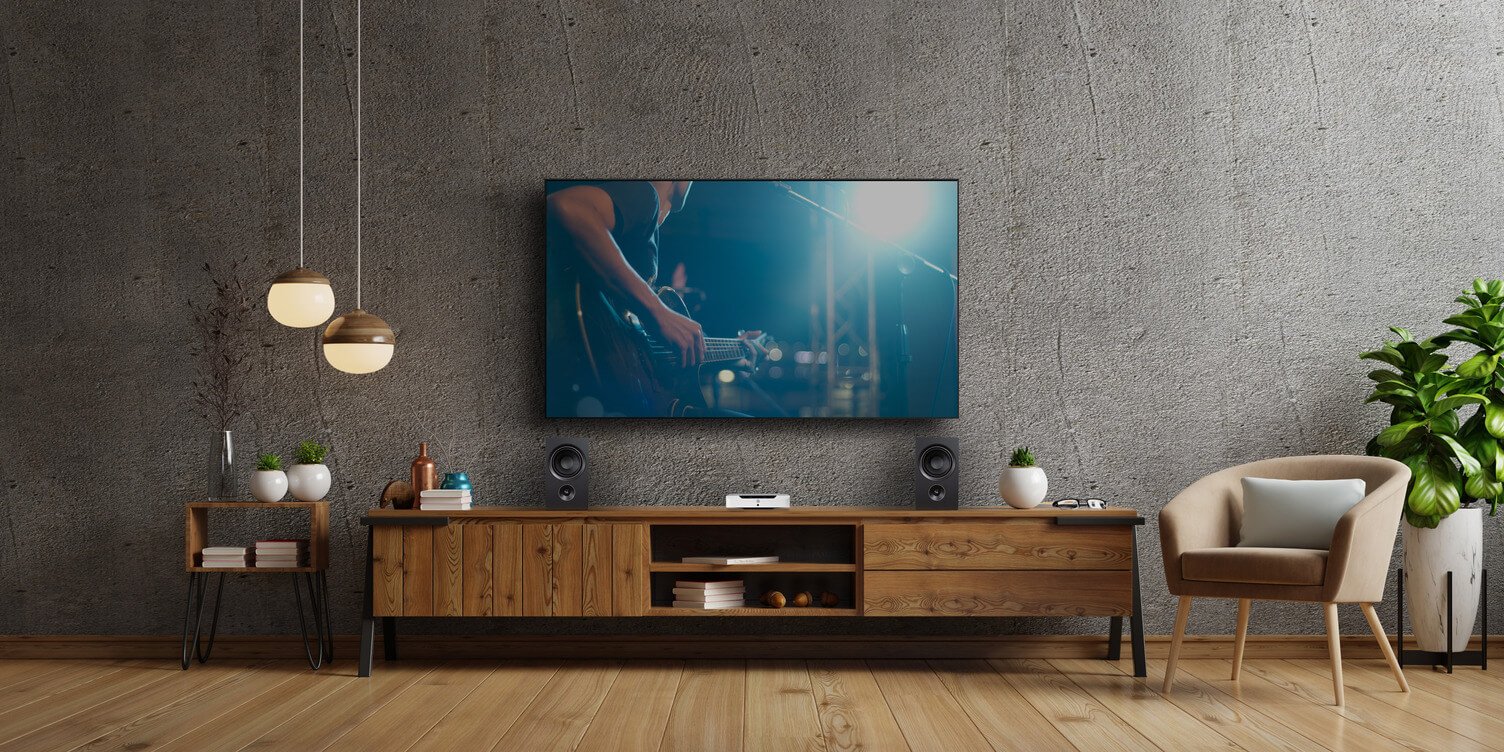 Wzmacniacz stereo z HDMI Powernode podłączony do telewizora, obok głośniki. Na ekranie widać mężczyznę trzymającego gitarę